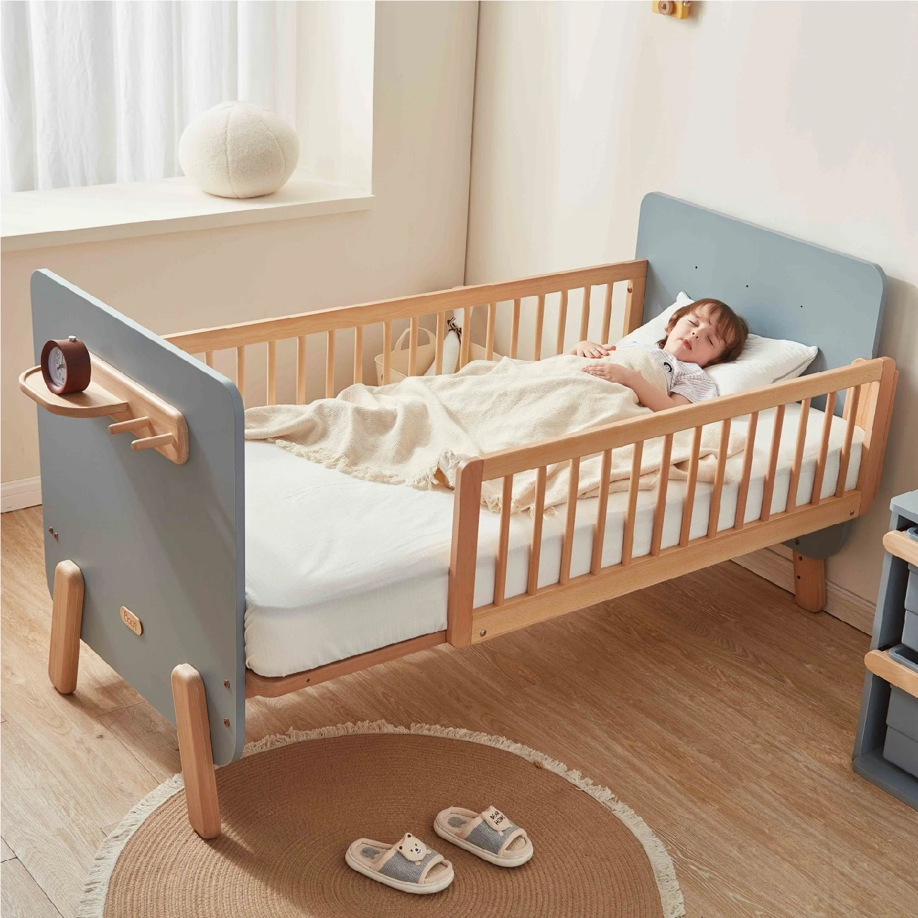Wooden convertible toddler bed with blue-grey headboard, side guardrails, and an attached shelf, with a child sleeping inside" Wooden convertible toddler bed with blue-grey headboard, side guardrails, and an attached shelf, with a child sleeping inside"