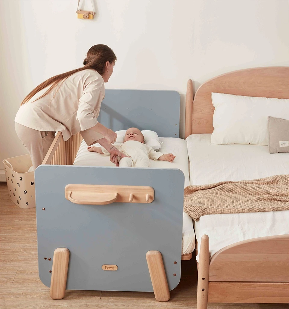 Parent leaning over a wooden bedside co-sleeper bed attached to an adult bed, settling a newborn Parent leaning over a wooden bedside co-sleeper bed attached to an adult bed, settling a newborn