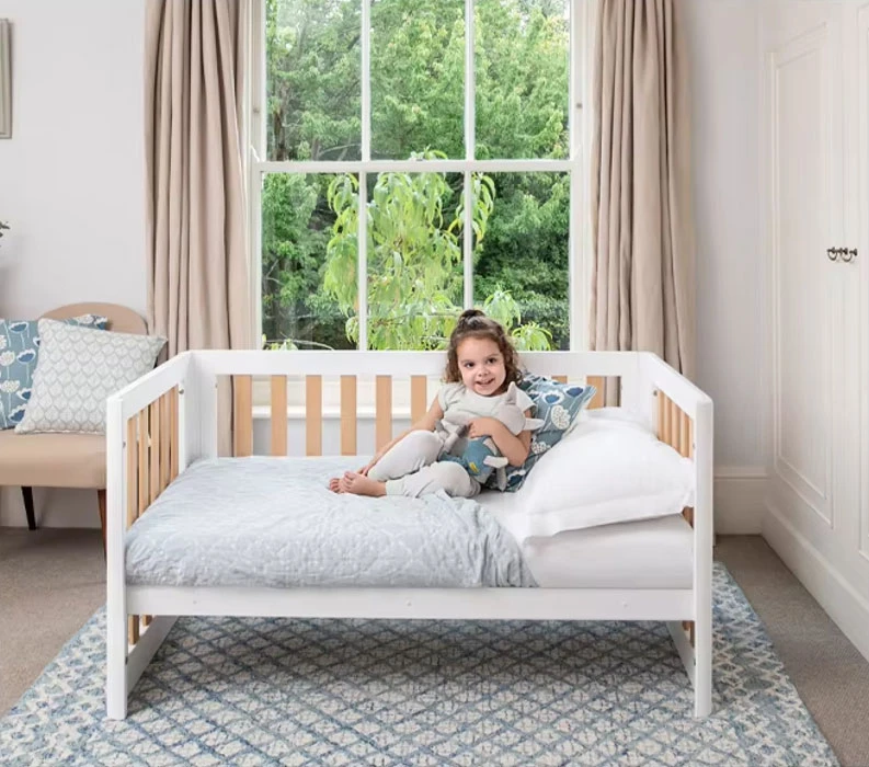 Smiling toddler sitting on a white wooden cot converted into a toddler bed, with natural slats behind her in a bedroom Smiling toddler sitting on a white wooden cot converted into a toddler bed, with natural slats behind her in a bedroom