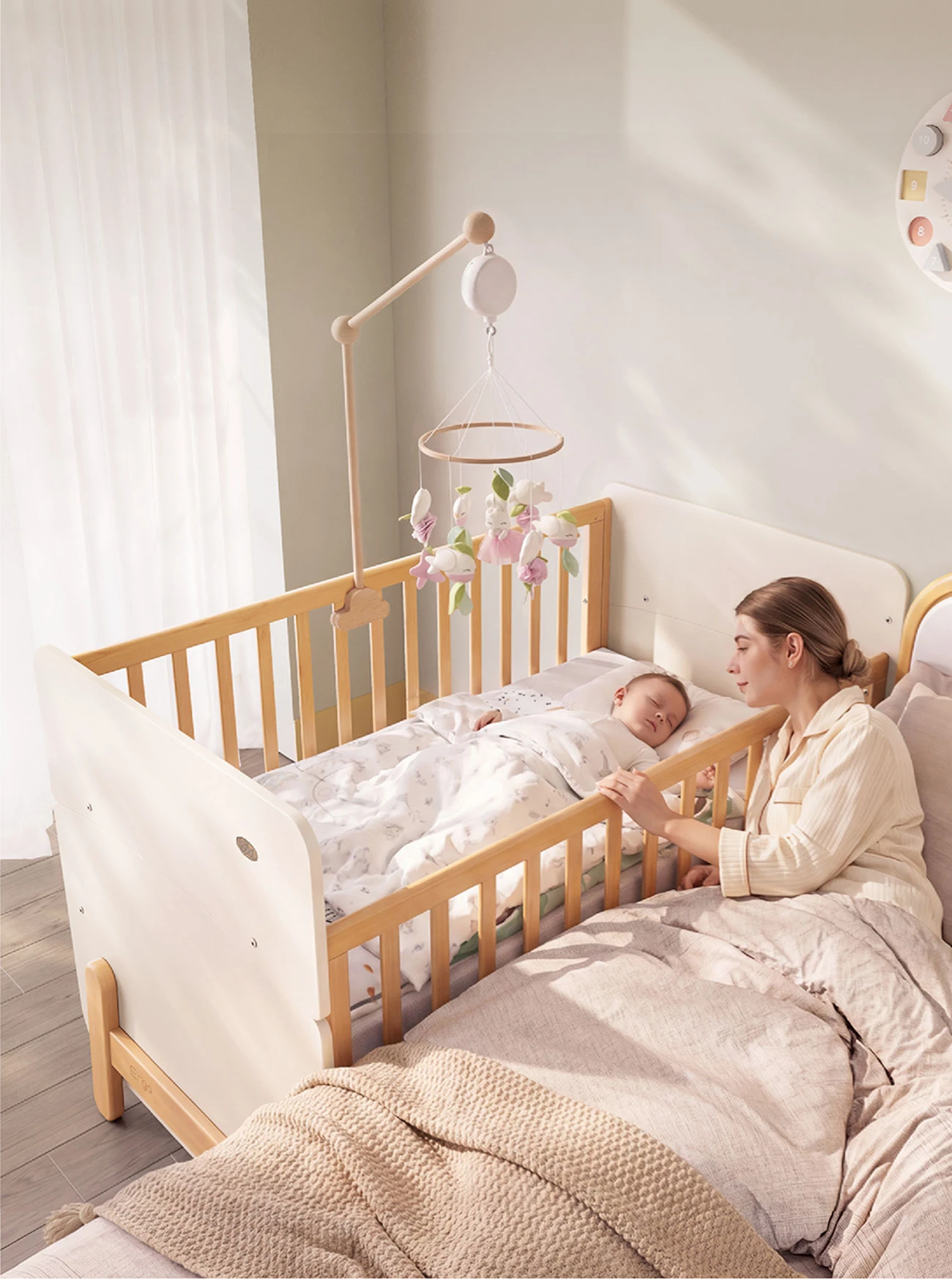Baby sleeping in a wooden cot with white end panels placed next to a parent's bed, with a mother resting beside it Baby sleeping in a wooden cot with white end panels placed next to a parent's bed, with a mother resting beside it