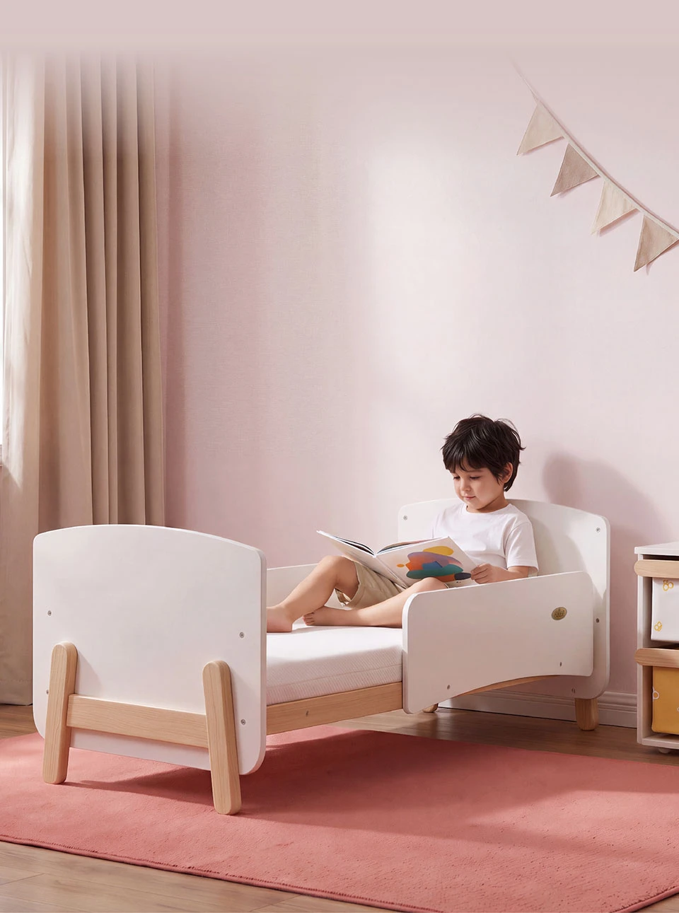 Boy reading a book on a low toddler bed with white end panels and timber feet, in a pink bedroom Boy reading a book on a low toddler bed with white end panels and timber feet, in a pink bedroom